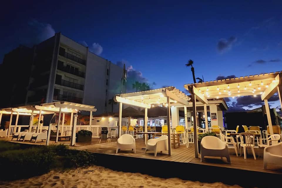 Illuminated beachfront deck pavilions at twilight with lounge seating, wooden pergolas, and sand