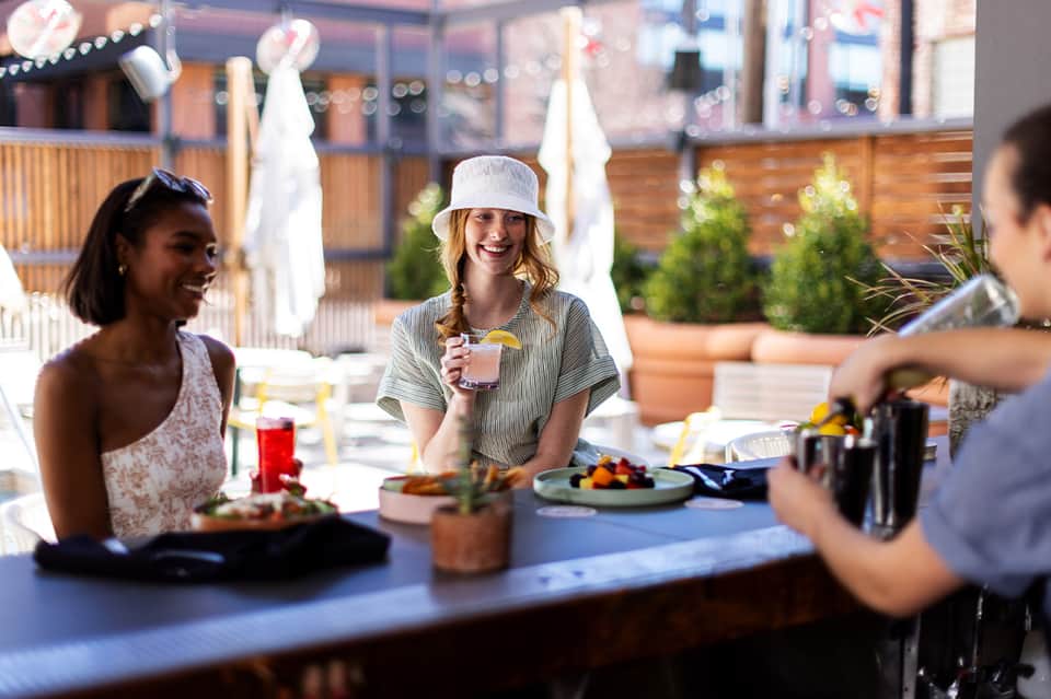 Rooftop dining area with guests enjoying meals, white umbrellas, string lights, and city views