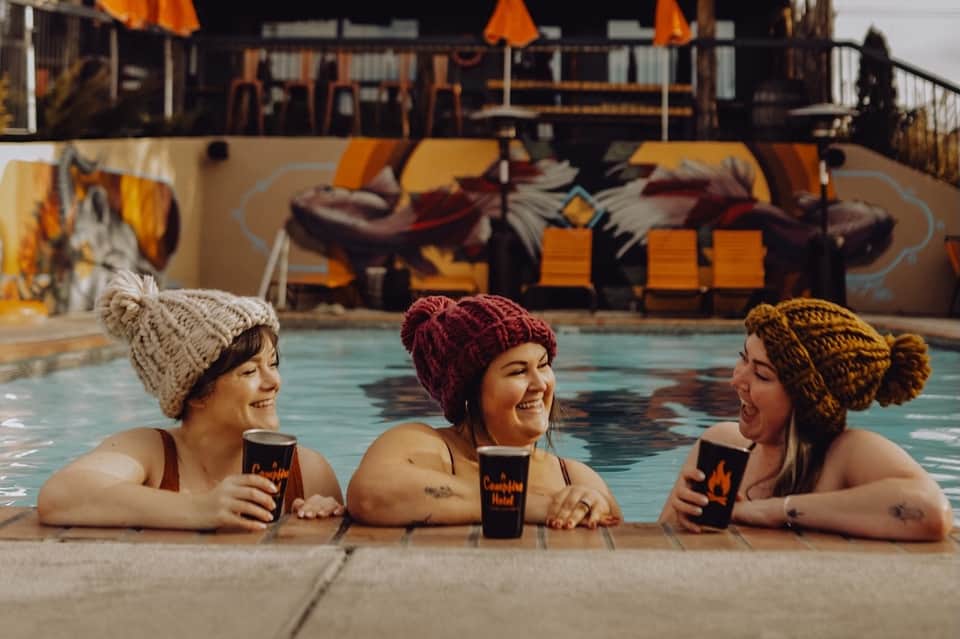 Three women in winter hats enjoying hot drinks at an outdoor heated pool with lounge chairs