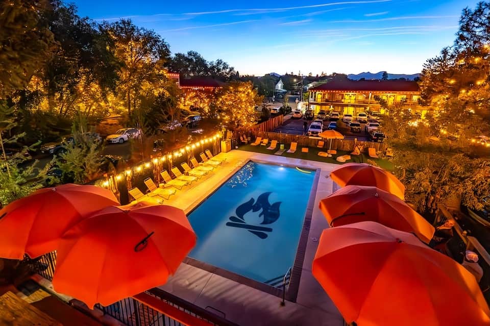 Illuminated resort pool at night with red umbrellas, lounge chairs, and glowing landscape lighting