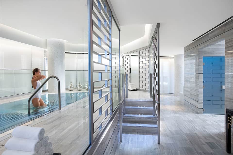 Modern indoor swimming pool with natural lighting, columns, person at pool edge, spa-like design aesthetic