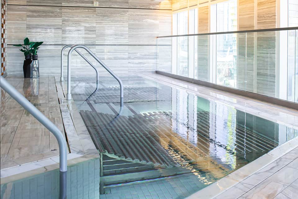 Indoor swimming pool with glass railings, lounge chairs, and natural light from windows