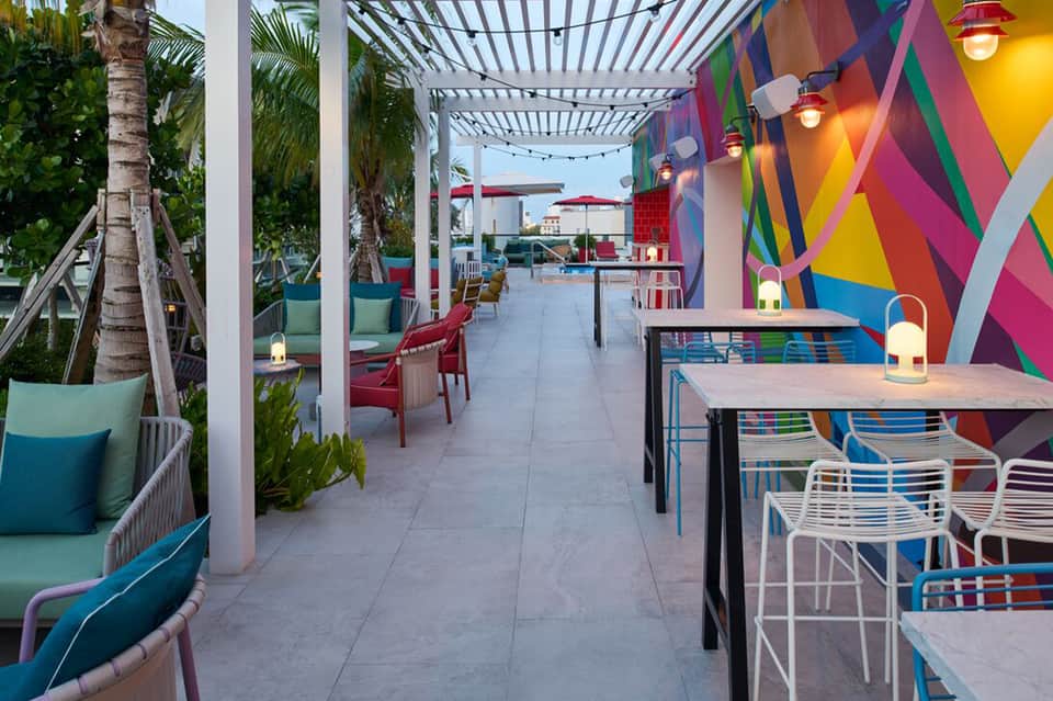 Rooftop lounge with colorful geometric wall art, pergola, dining tables, and tropical plants