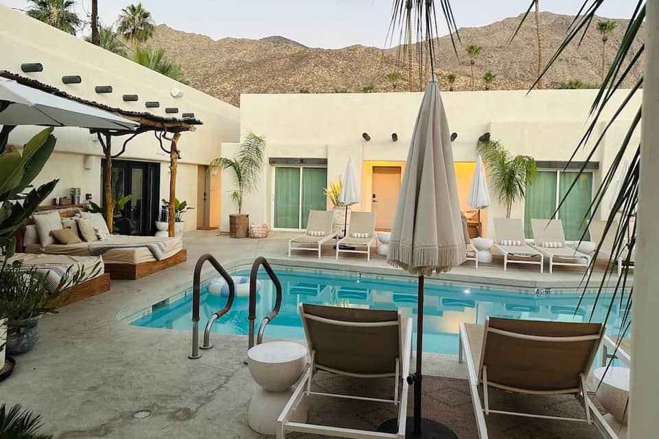 Modern resort courtyard with rectangular pool, lounge chairs, umbrellas, daybed seating, and white buildings against desert mountain backdrop