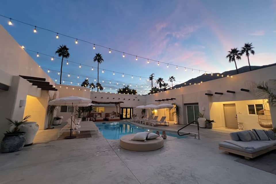 Resort courtyard at dusk with illuminated pool, fire pit, loungers, string lights, and palm trees