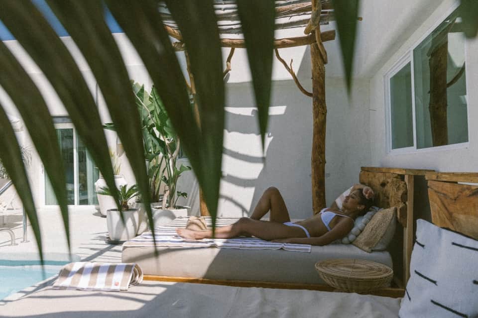 Woman relaxing on outdoor daybed with palm fronds, white pillows, and natural sunlight