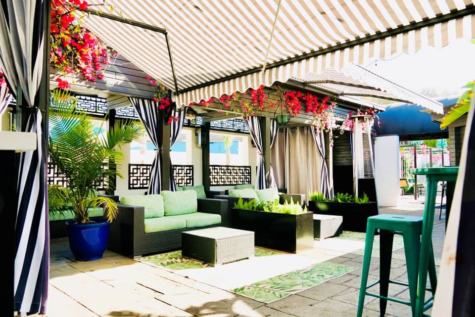 Covered outdoor patio with striped awning, hanging flowers, black furniture, and tropical plants