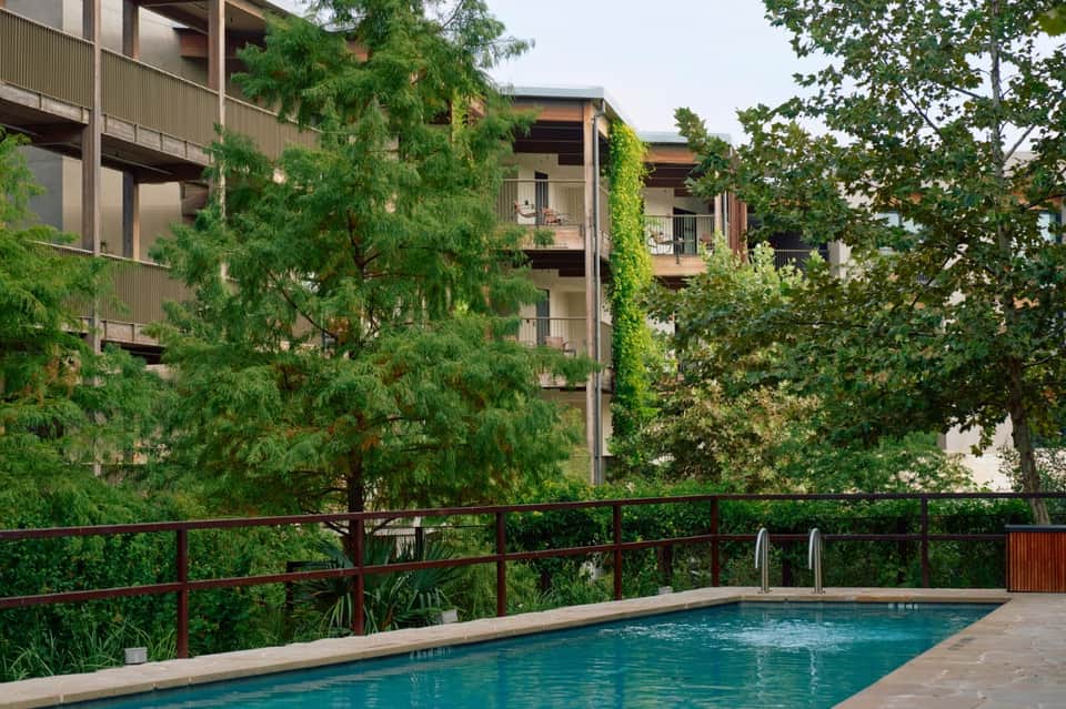 Resort pool surrounded by lush green trees and tropical vegetation with metal railing and apartments