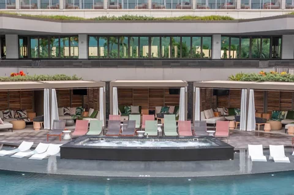 Resort pool deck with multi-colored loungers, cabanas with curtains, hot tub, and modern building