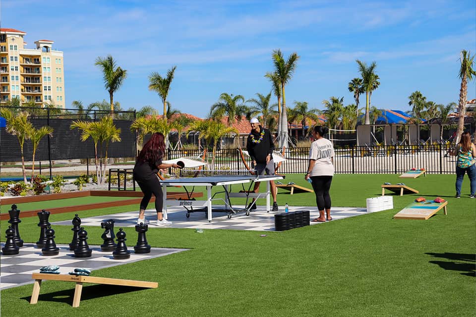 Outdoor recreation area with lawn games, ping pong table, giant chess set, and palm trees with buildings