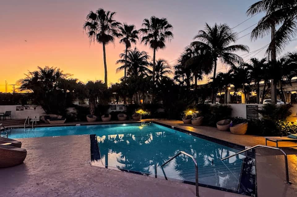 Resort swimming pool at dusk with palm trees, ambient landscape lighting, lounge seating, sunset sky