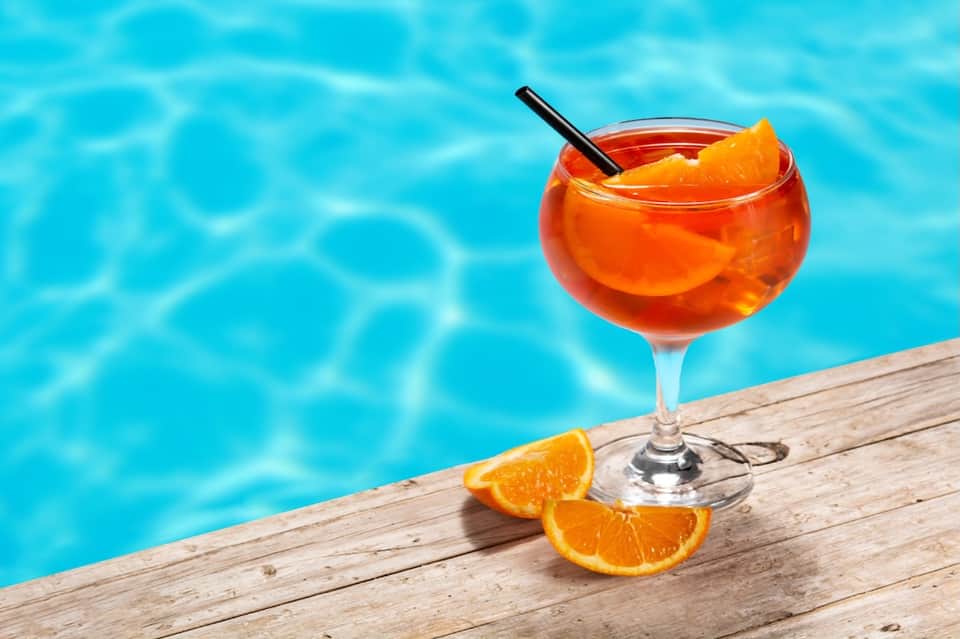 Orange cocktail with citrus slices on poolside wooden deck with sparkling blue water in background