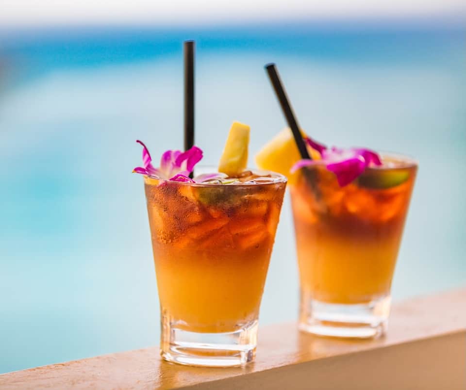 Two tropical cocktails with orange liquid, pineapple garnish, and blue ocean backdrop