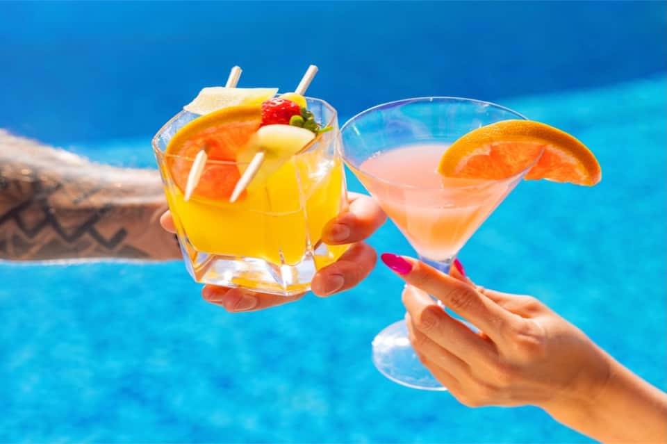 Hands holding colorful tropical cocktails over bright blue swimming pool water