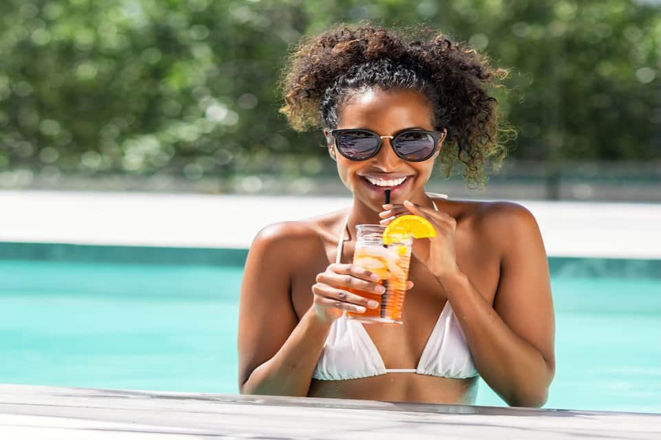Woman enjoying tropical cocktail poolside wearing sunglasses and white bikini