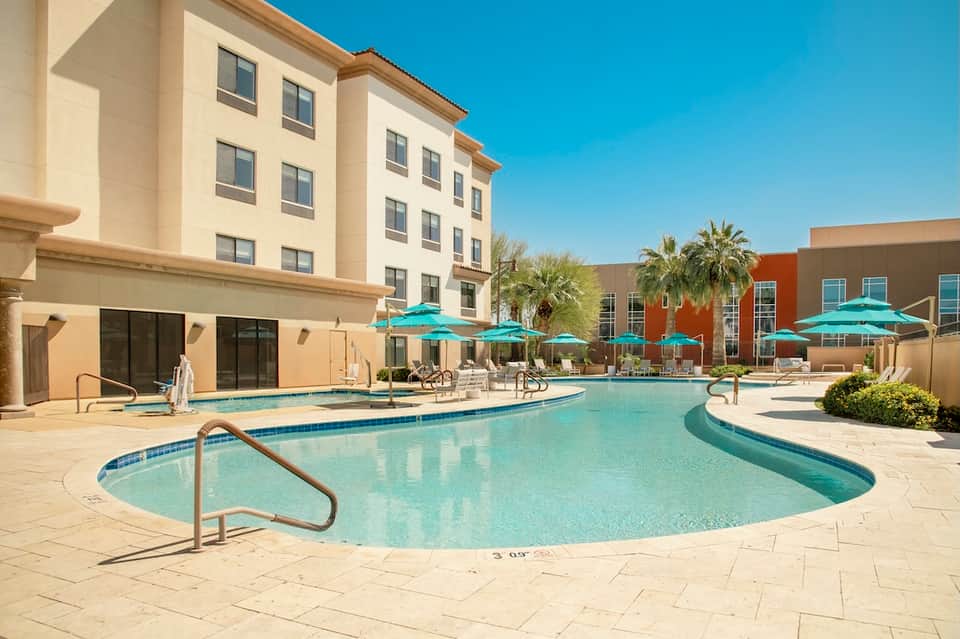 Resort pool area with turquoise water, white umbrellas, palm trees, and beige building facade