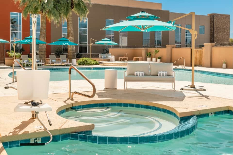 Resort pool area with turquoise umbrellas, lounge chairs, spa tub, and modern building