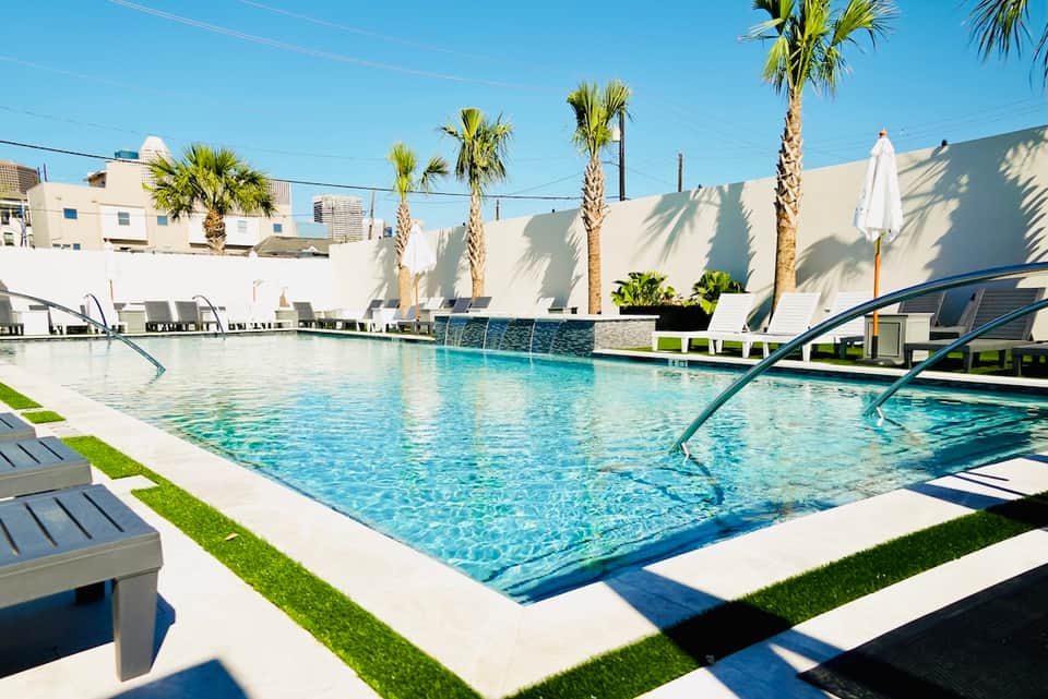 Resort swimming pool with clear blue water, white loungers, palm trees, and modern architecture