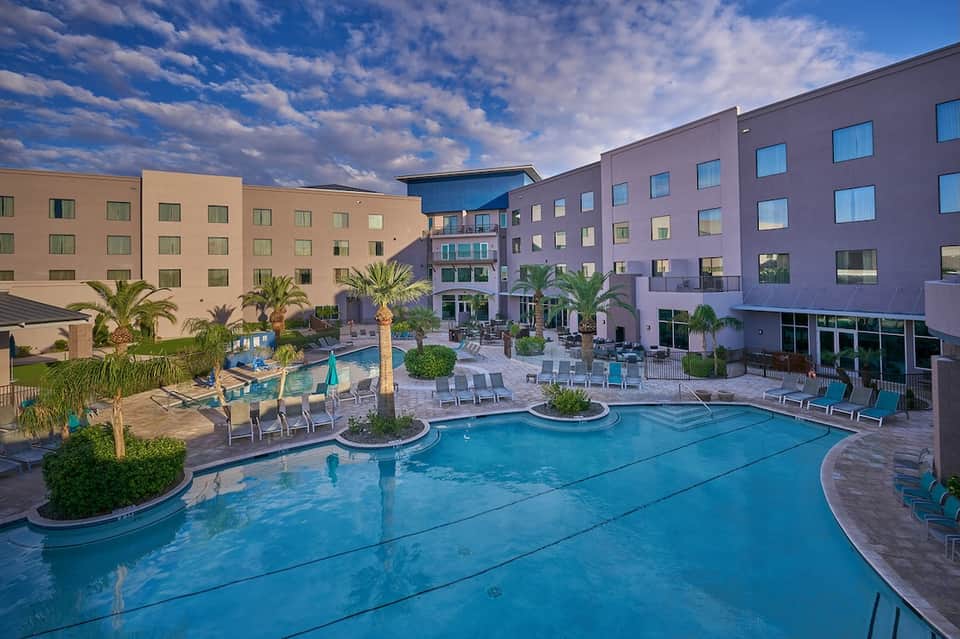 Large resort pool with lap lanes, multiple loungers, palm trees, and contemporary hotel buildings