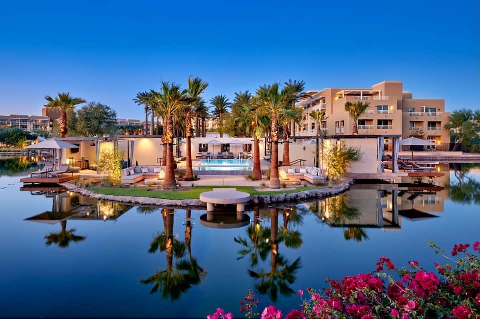 Waterfront resort with palm trees, pool area, modern buildings reflected in calm lagoon