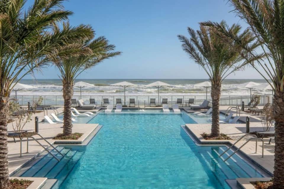Beachfront resort pool with palm trees, white lounge chairs, ocean waves, and clear sky