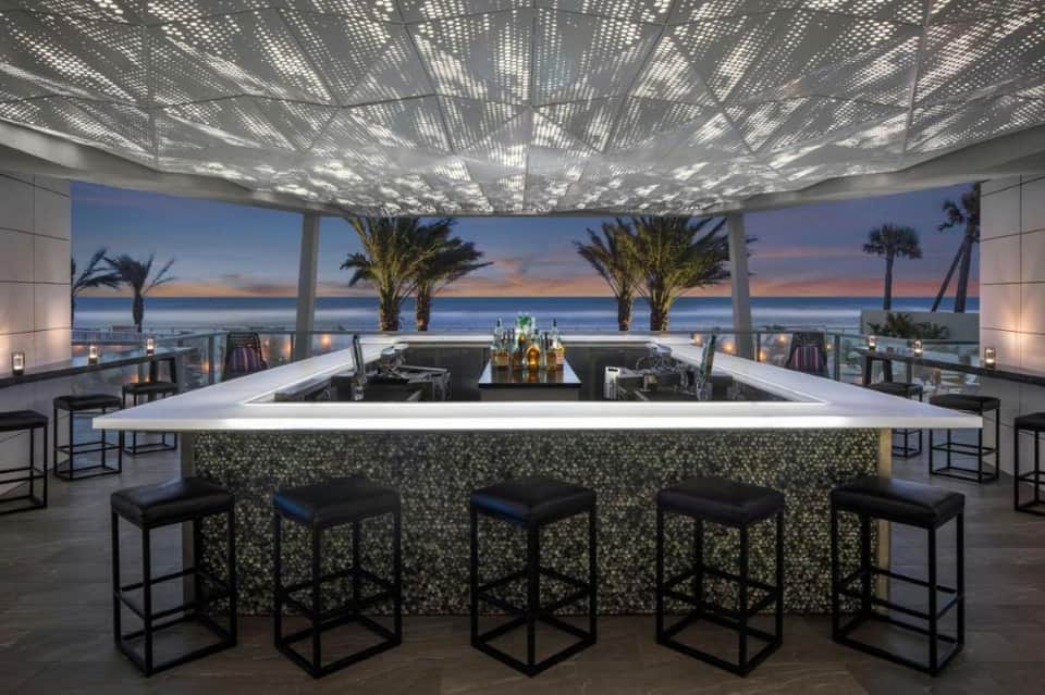 Rooftop bar with panoramic ocean and sunset views, perforated ceiling, modern counter seating, and palm trees