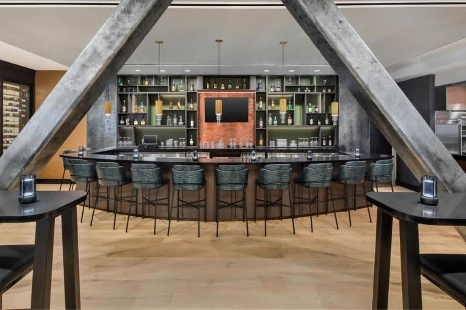 Contemporary hotel bar with curved seating, large concrete support beams, extensive bottle display, and ambient lighting