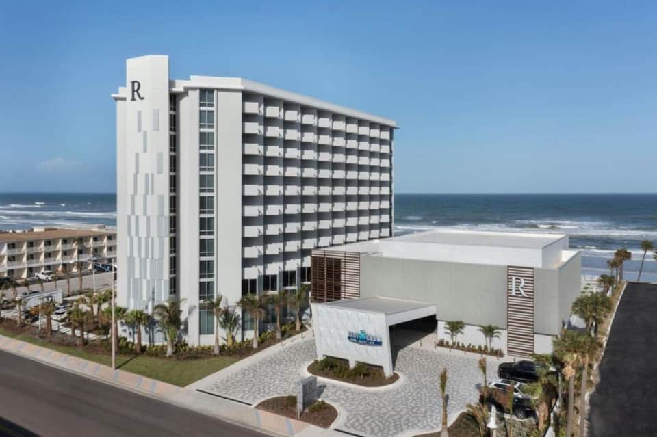 Beachfront resort building with white tower, modern architecture, ocean view, and palm trees