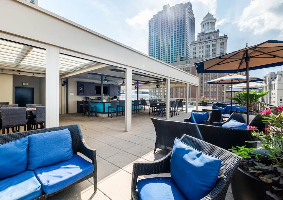Rooftop terrace with modern lounge chairs featuring blue cushions under extended pergola with city skyline views and blue bar