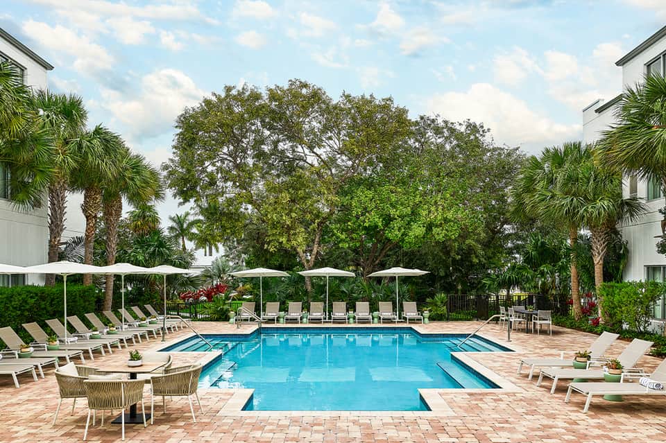 Resort-style pool area with lounge chairs, white umbrellas, palm trees, and manicured landscaping
