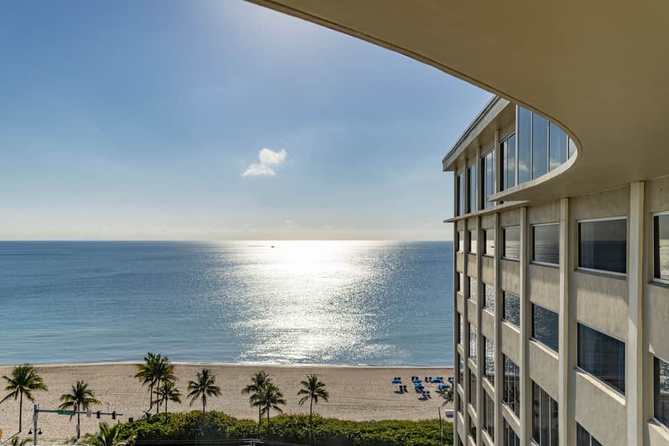 Beachfront balcony view with palm trees, sandy shore, and sparkling ocean under blue sky