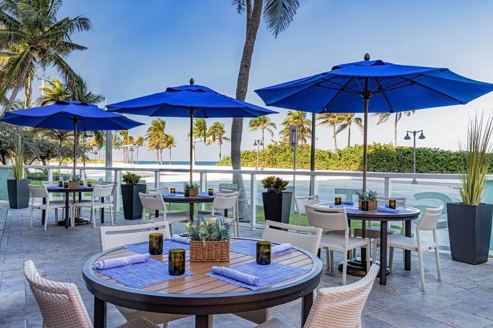 Waterfront dining area with blue umbrellas, white wicker chairs, and palm trees