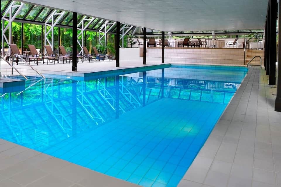 Indoor pool facility with glass ceiling, turquoise water, white lane markers, metal structural framework, and lounge area.