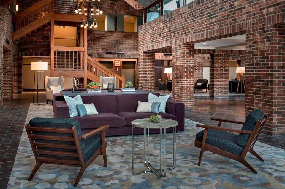 Spacious lobby lounge with exposed brick, purple sofa, wooden ceiling beams, and mid-century chairs