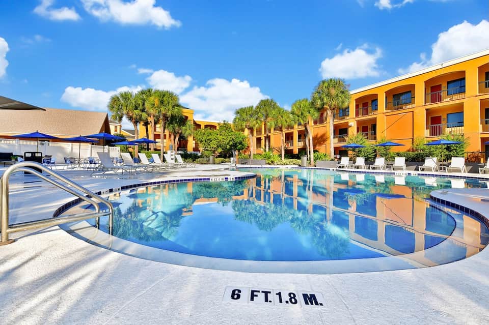 Resort pool with depth marking, surrounded by yellow buildings, palm trees, and lounge seating areas