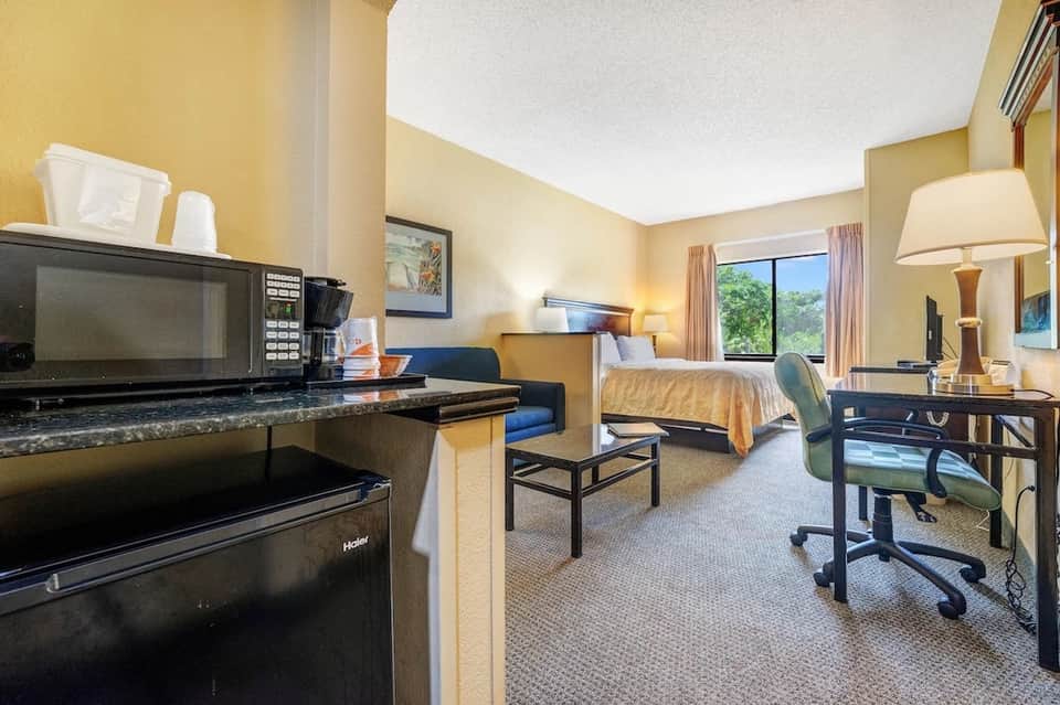 Hotel room with yellow walls, queen bed, work desk, microwave, and large window overlooking grounds
