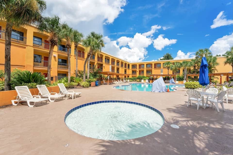 Resort courtyard with yellow building, palm trees, swimming pool, and white lounge chairs on sunny day
