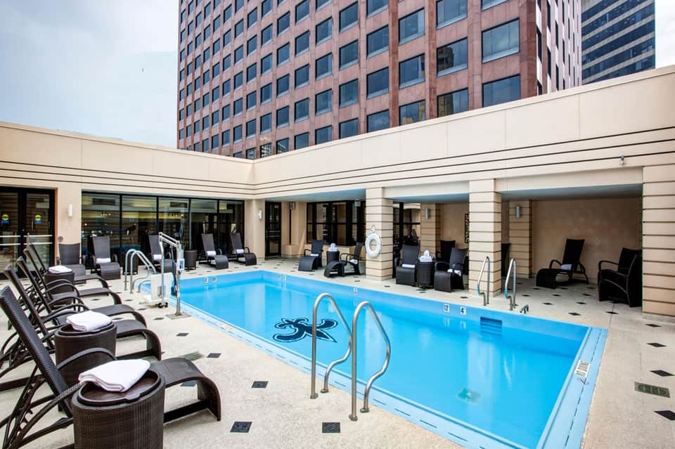 Indoor hotel pool with bright blue water, lounge chairs, dark wicker seating, and tall brick building backdrop.