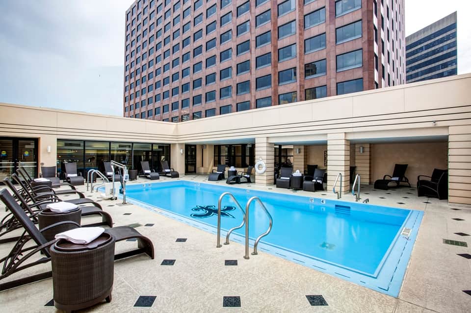 Indoor hotel pool surrounded by black lounge chairs with white towels and tall brick buildings overhead