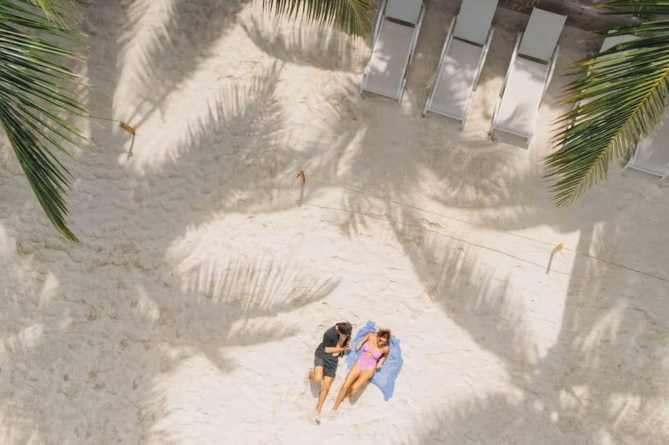 Overhead view of two guests relaxing on white sand beach between palm fronds