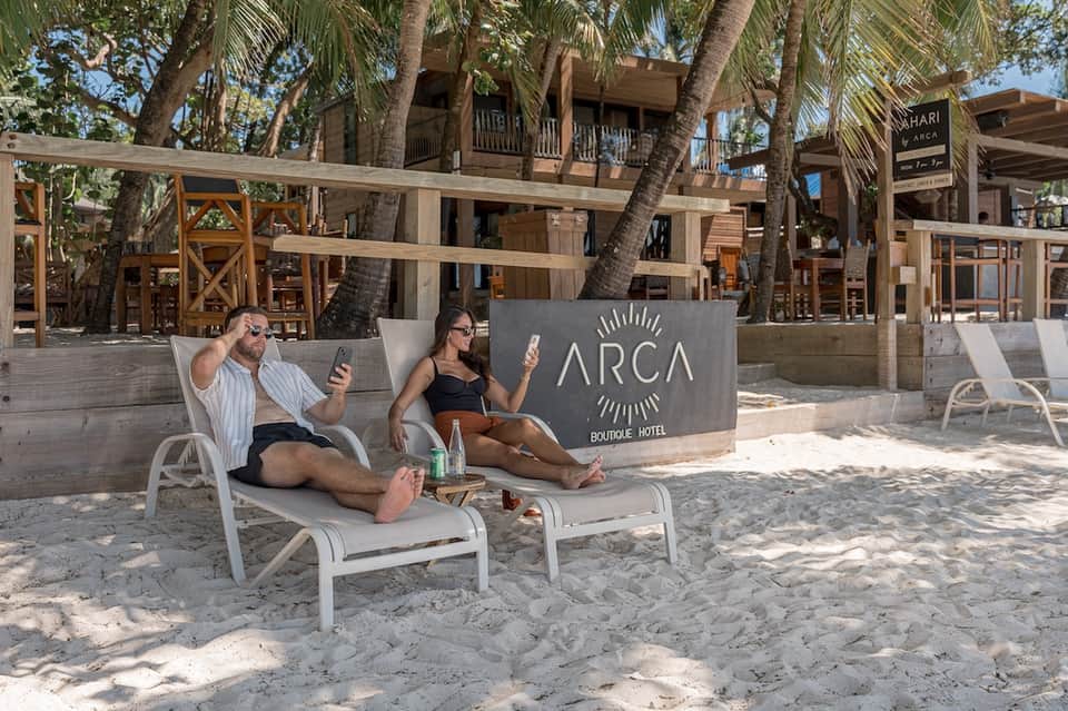 Beachfront lounge area with palm trees, ARCA Boutique Hotel signage, and guests relaxing on white loungers