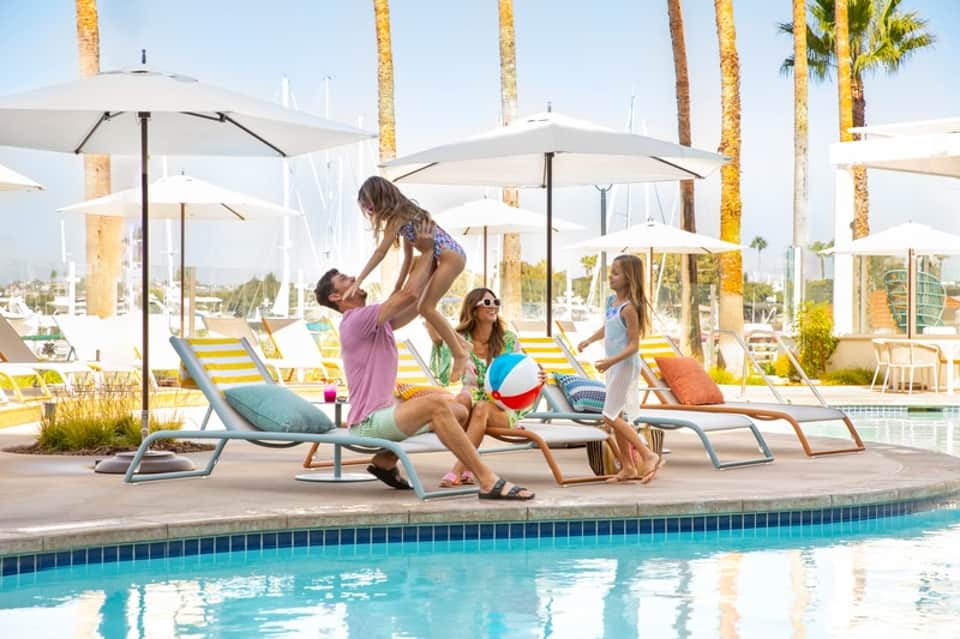 Resort poolside gathering with families, white umbrellas, lounge chairs, and palm trees