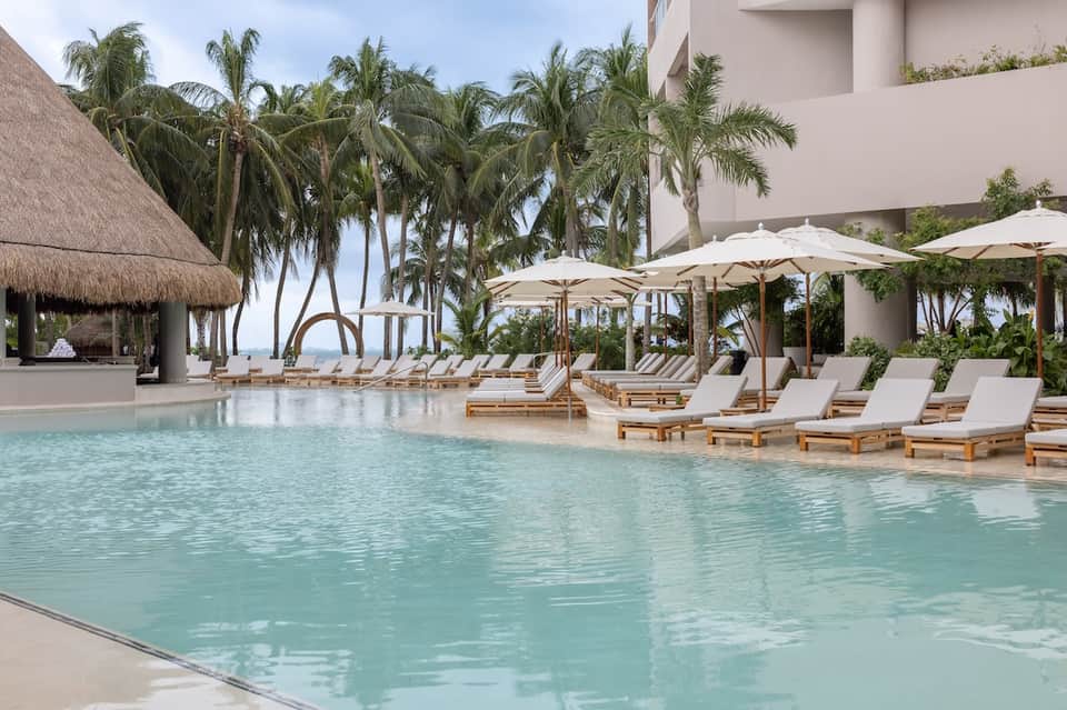 Beachfront resort pool with palm trees, lounge chairs, umbrellas, and tropical landscape views