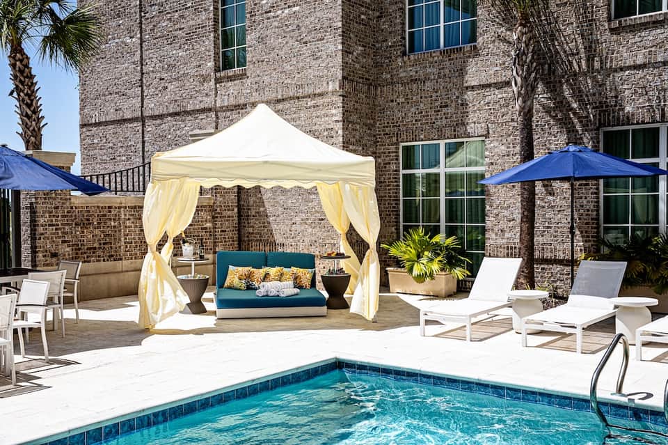 Poolside cabana with cream curtains and teal cushions beside brick building with blue umbrellas