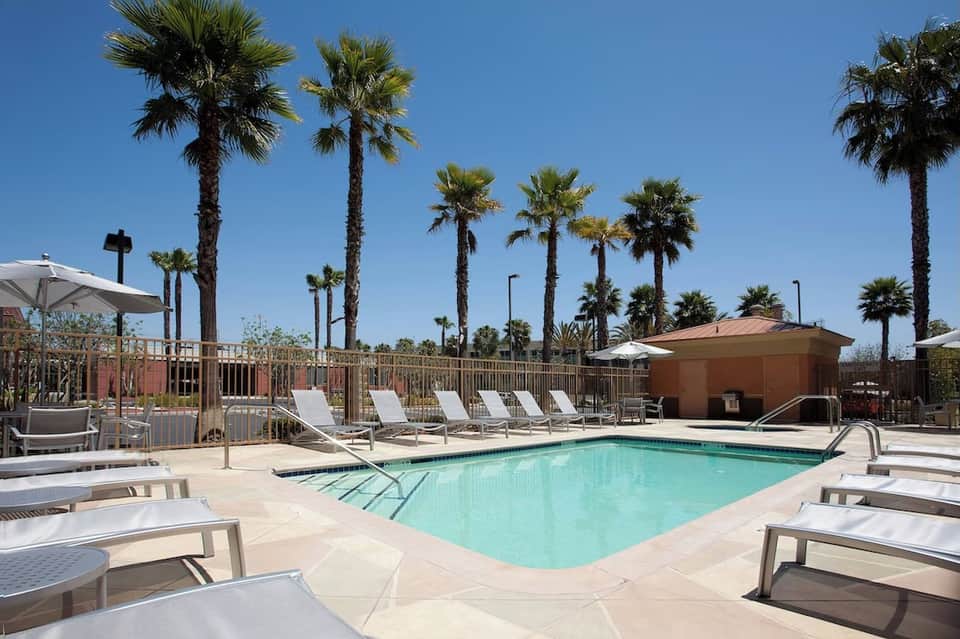 Resort pool surrounded by white loungers, palm trees, blue sky, and fenced recreational area