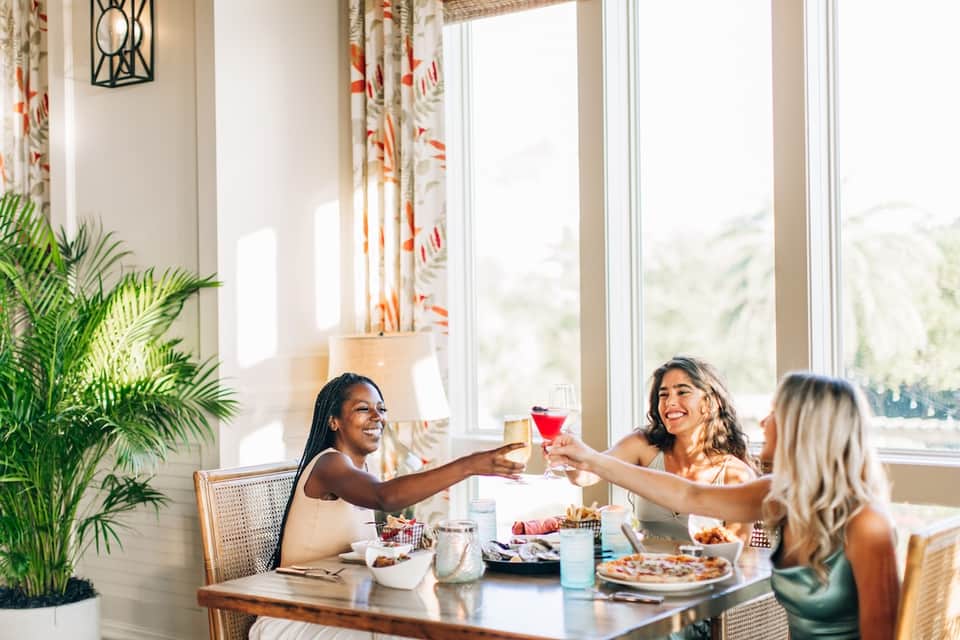 Three women dining indoors with tropical plants, cocktails, and ocean view through large windows