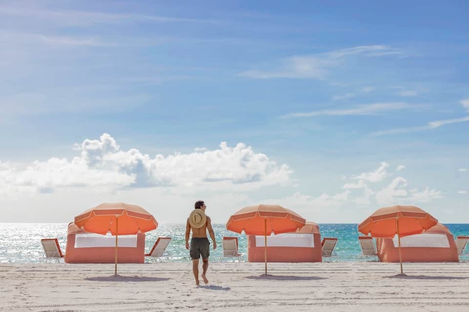 Beach scene with surfer walking toward orange and white beach umbrellas on sandy shore with ocean and clouds.