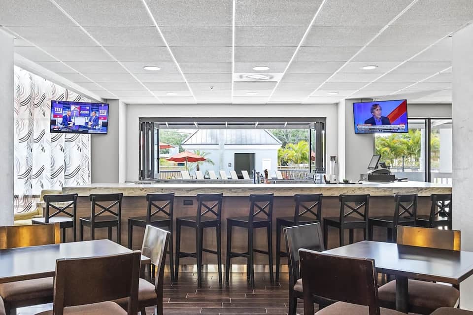 Hotel bar and lounge area with seating, counter, TV screens, and views to outdoor patio space