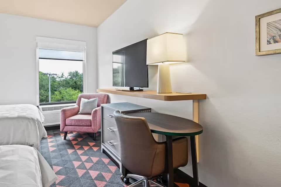 Hotel room with two beds, desk area, pink accent chair, and balcony with natural light