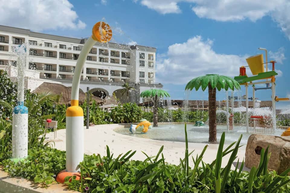 Playful splash pad area with water features, colorful props, palm trees, and residential building